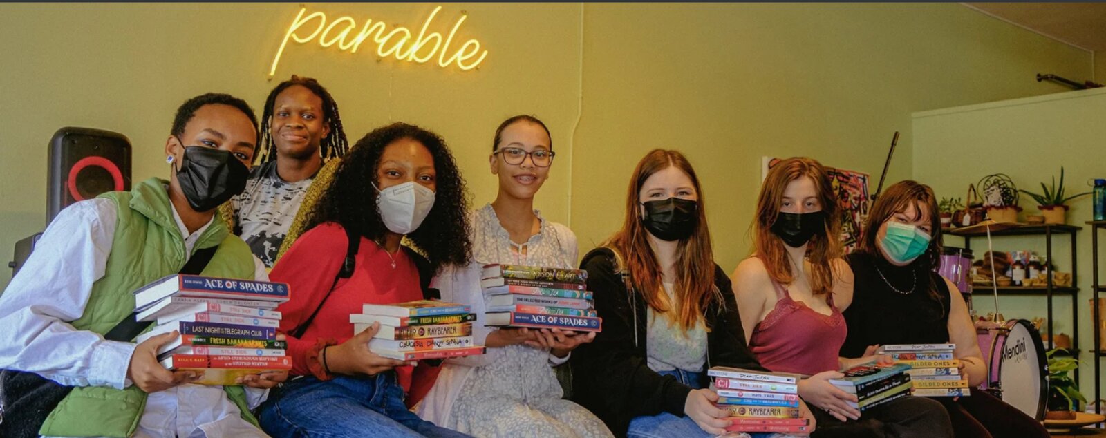 A group of students holding stacks of donated books in front of Parable's neon sign